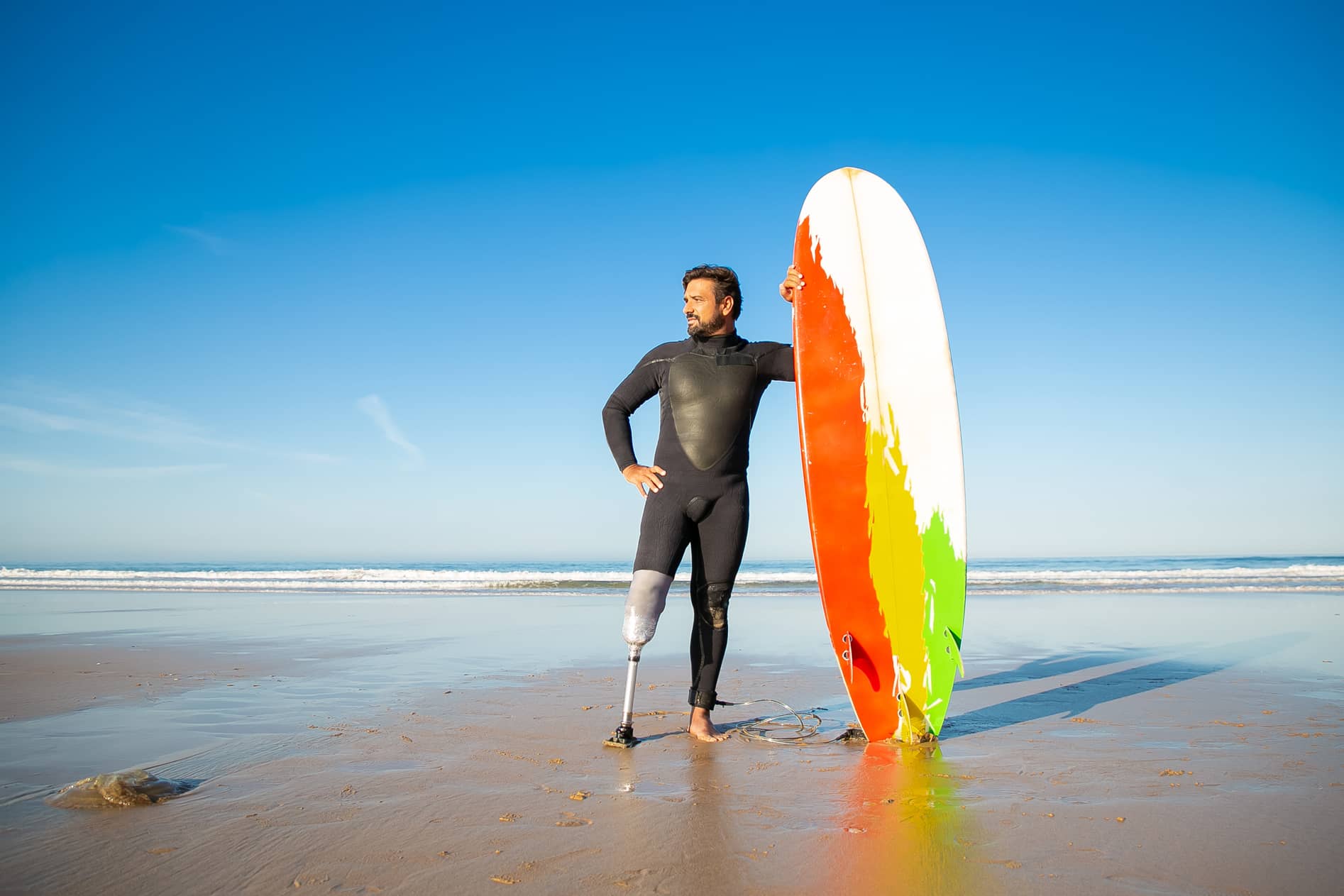  Surfeur avec prothèse de jambe posant avec sa planche sur la plage, discipline de glisse adaptée aux handicaps. 